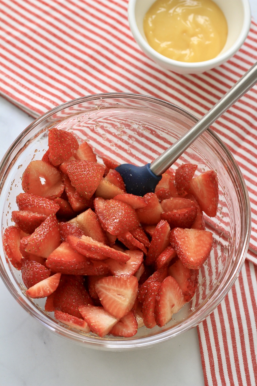 A glass bowl with slices of strawberry with a spatula and a white bowl with lemon curd in the top right on a white and red striped towel.