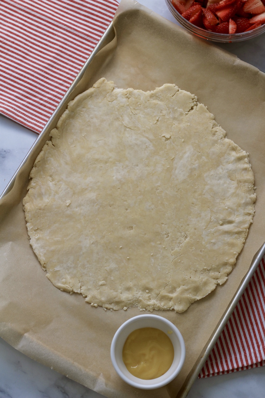 A rimmed baking sheet lined with parchment paper and a round of pie pastry with a white bowl in the bottom left corner.