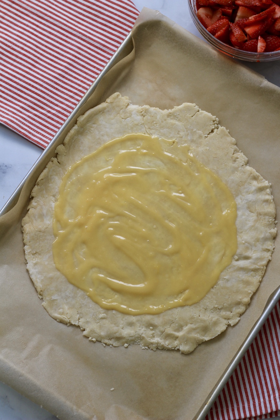 A rimmed baking sheet with parchment paper with a round pastry topped with lemon curd and a bowl of strawberries in the back.