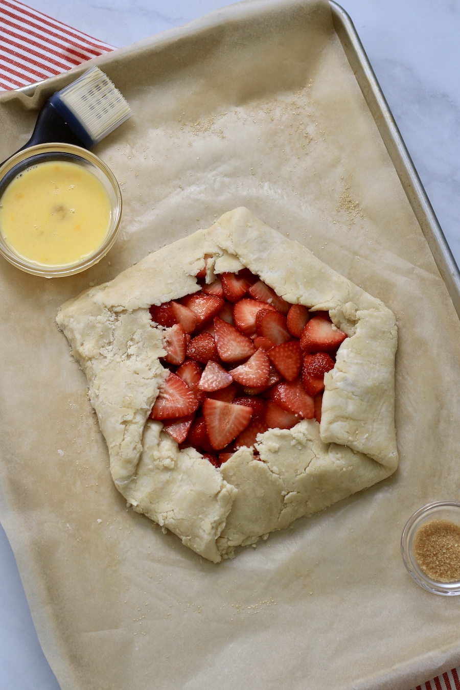 A rimmed baking sheet with unbaked strawberry lemon galette and a small bowl in the top left with a pastry brush.