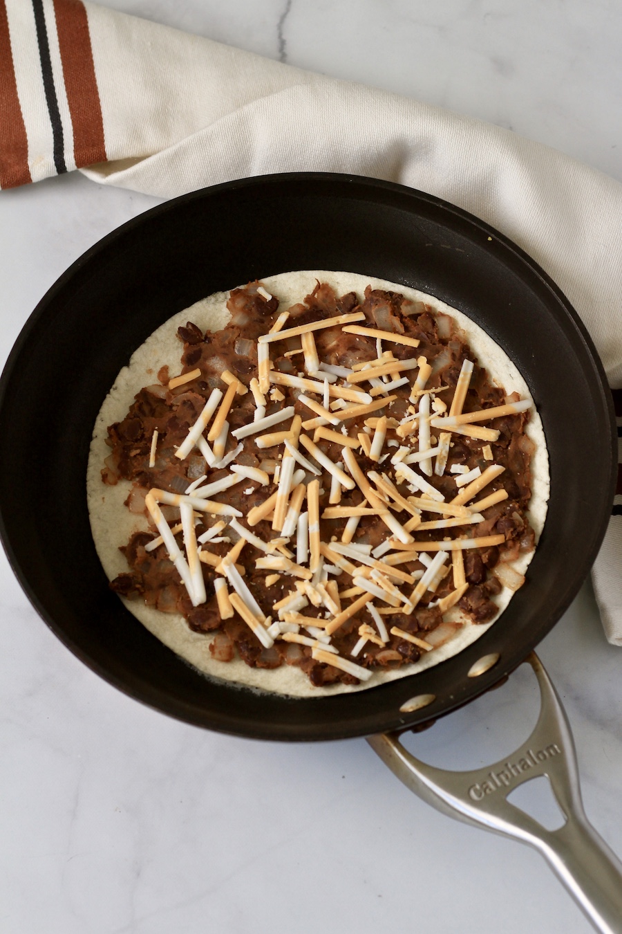 A skillet showing a tortilla with black bean filling and cheese shreds.