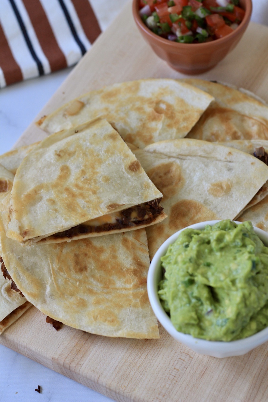 A wooden cutting board with wedges of vegan black bean and cheese quesadillas with a white bowl of guacamole to the right and an orange bowl of pico de gallo in the back.
