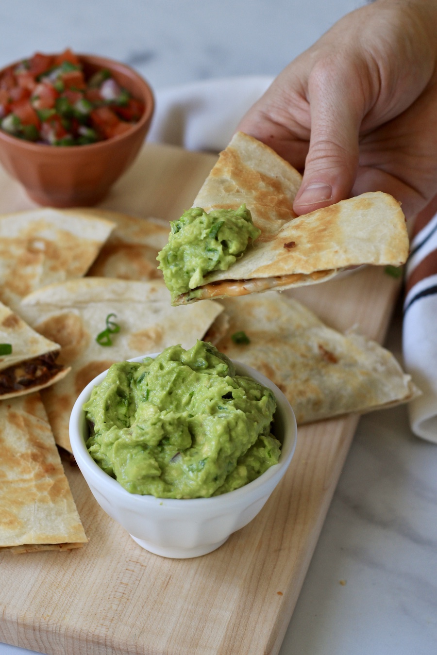 A hand with a wedge of quesadilla dipped in guacamole with a white bowl of guacamole on the wooden cutting board.