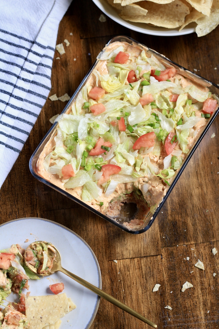 A top down photo of vegan 7 layer dip in a glass dish with a plate in the front left with a gold spoon and a bowl of tortilla chips in the back.
