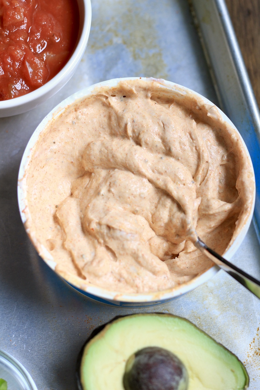 A white bowl with the sour cream layer with a spoon in front of an avocado.