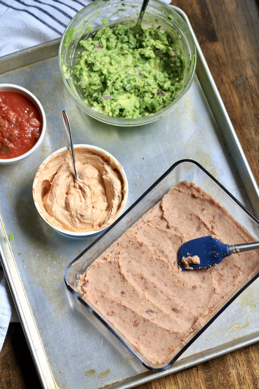 The refried beans in a glass dish with a spatula in front of a bowl of the sour cream mixture, a guacamole bowl, and salsa bowl.