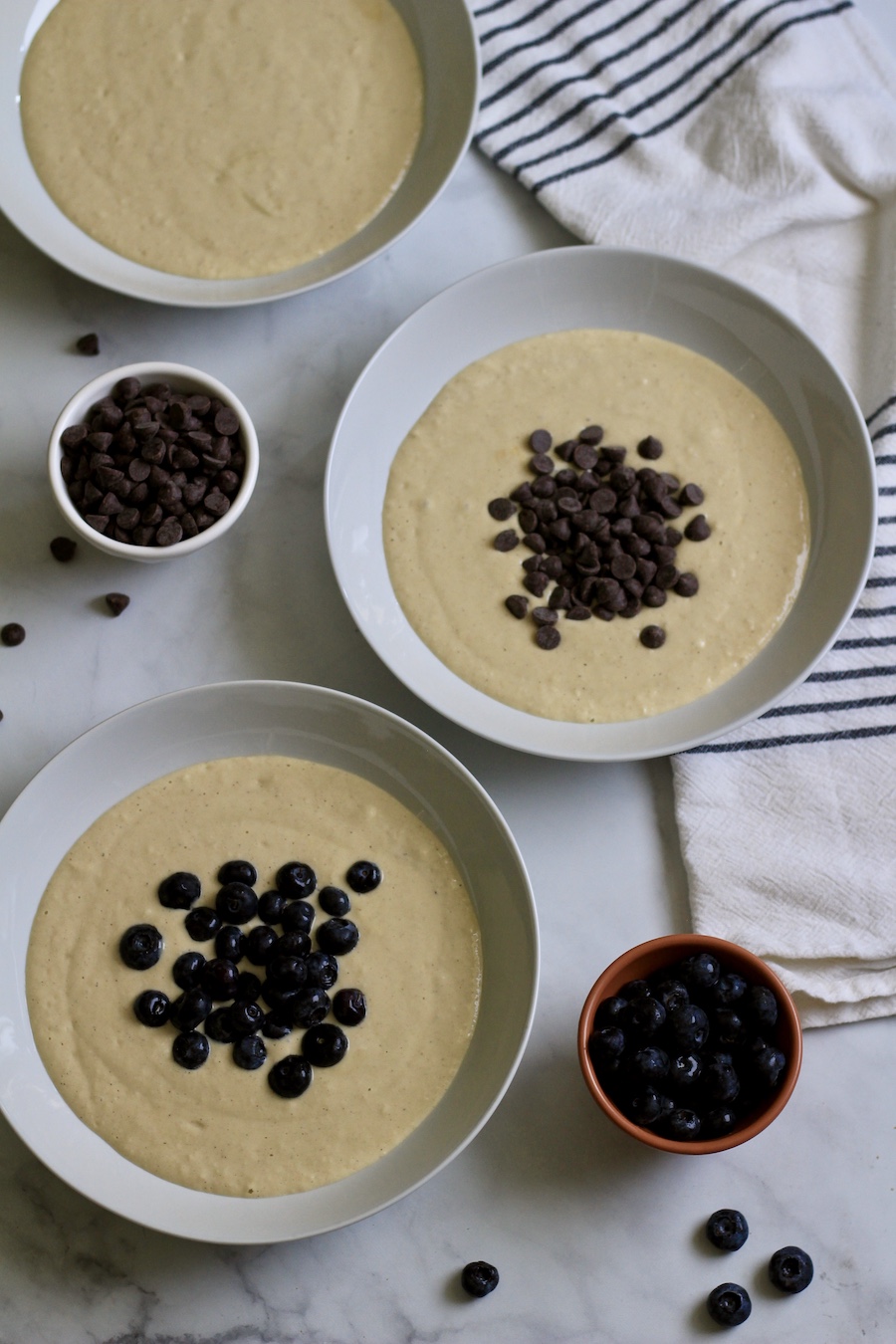 Three bowls of pancake batter the one at the top back is plain the one in the middle right has chocolate chips and the one in the bottom left has blueberries.