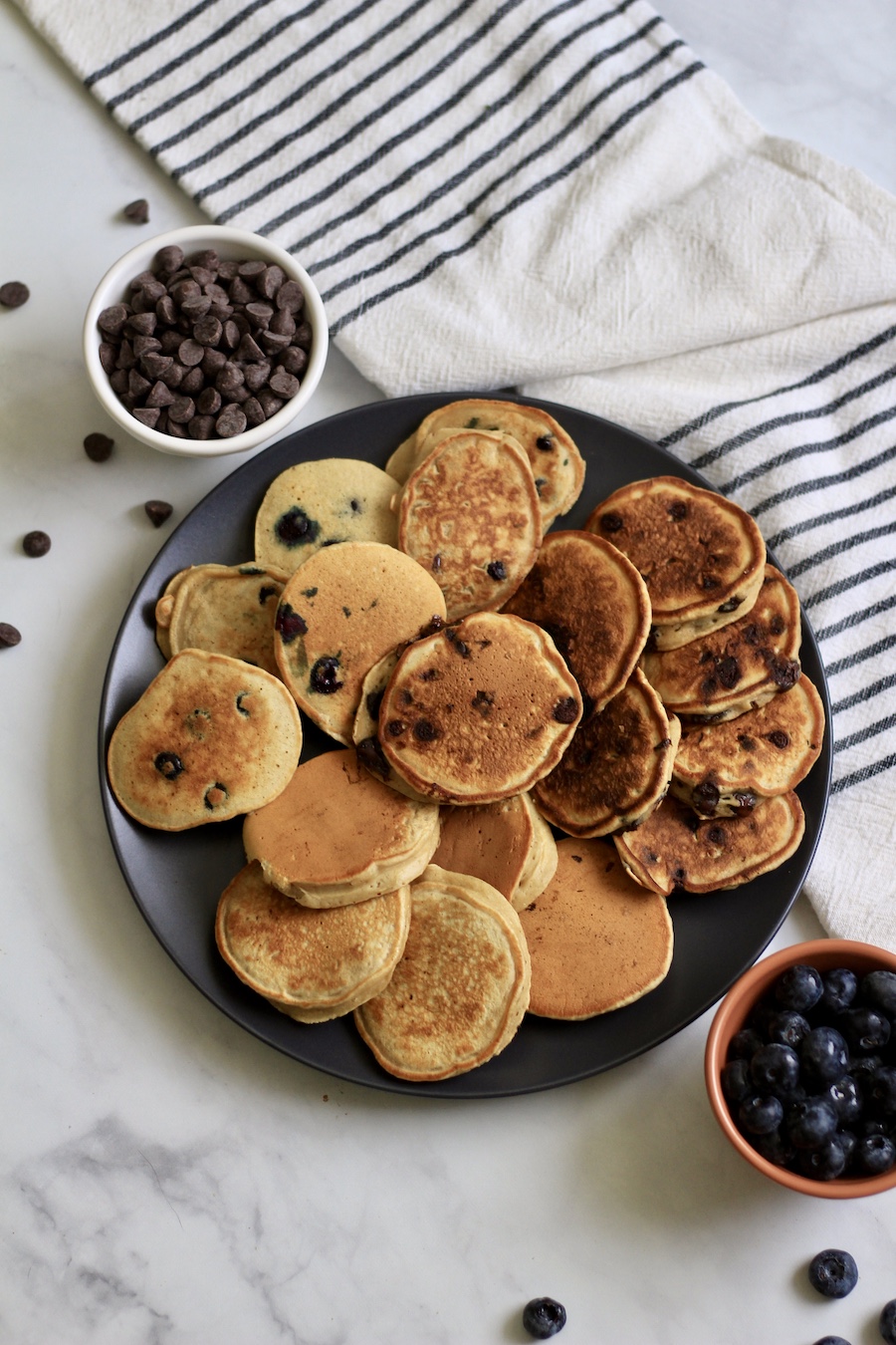 A blue plate with mini-pancakes and a bowl of chocolate chips in the top right and a bowl of blueberries in the bottom right.