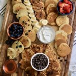 A wooden cutting board with mini-buttermilk pancakes and additional toppings in bowls.