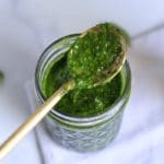 Square photo of a glass jar with a gold spoon dipped in spinach pesto.