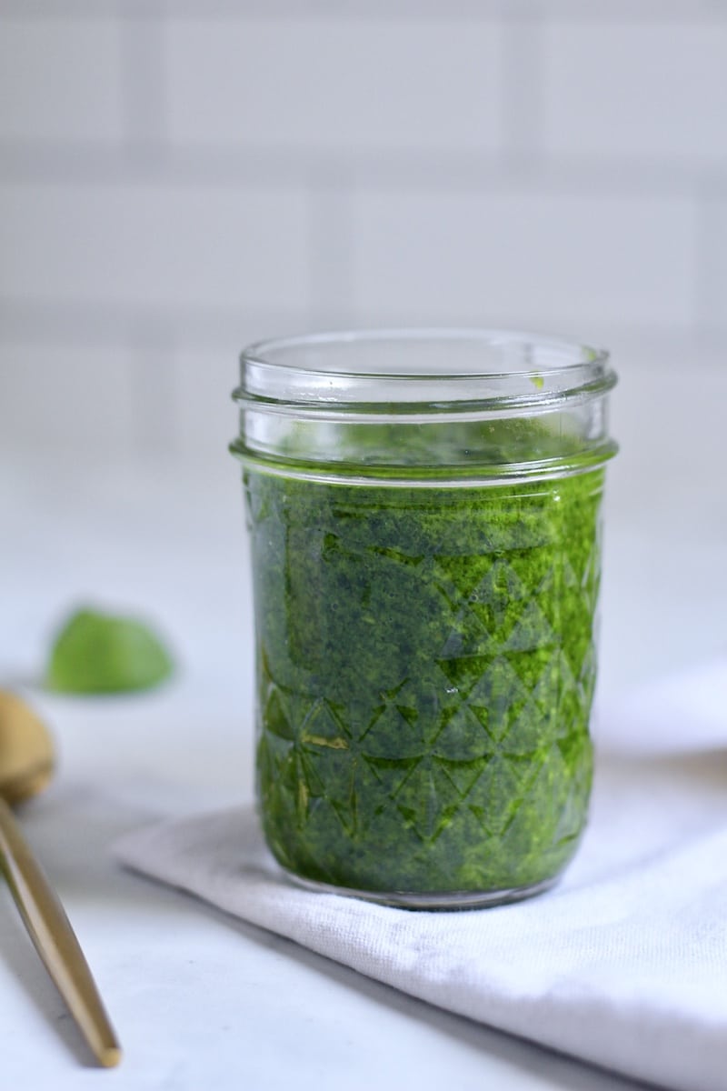 A glass jar of spinach pesto on a white towel with a gold spoon to the left.