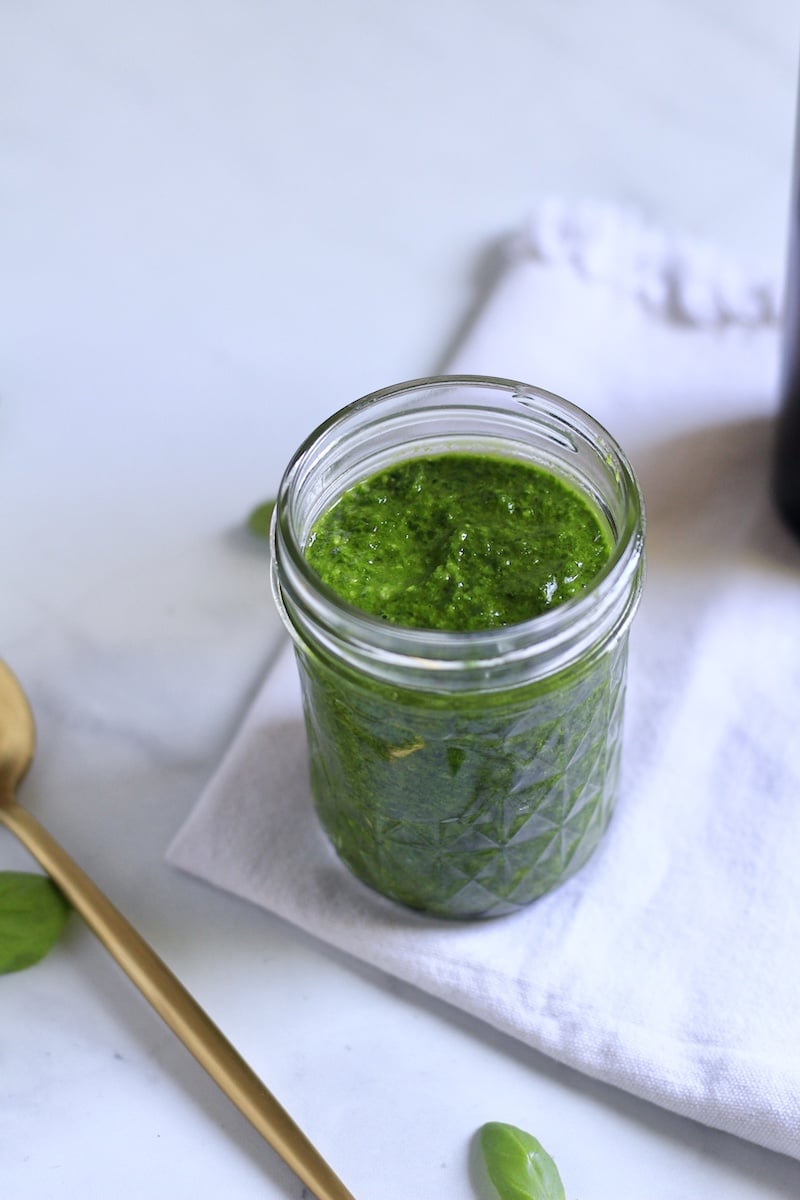 Spinach pesto in a glass jar on a white towel with a golden spoon to the left.