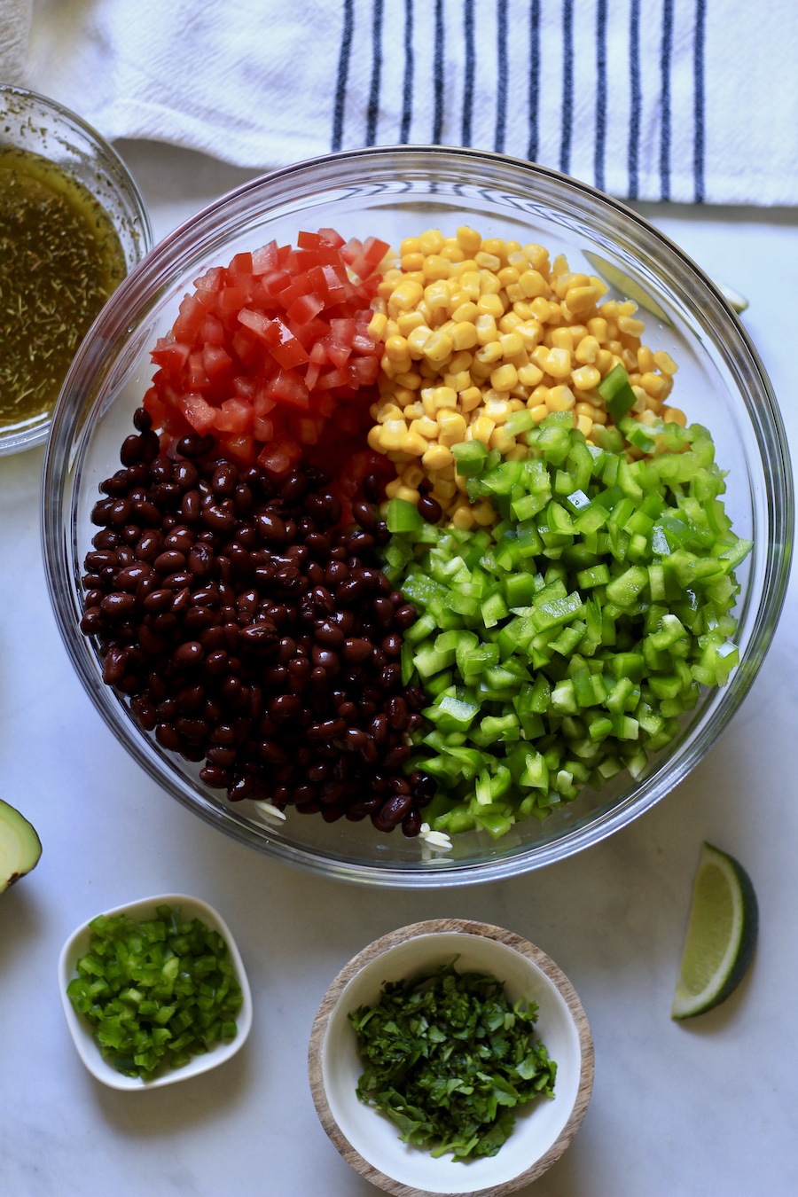 A glass bowl with the ingredients for texas caviar pasta salad with additional garnishes in small bowls in front of the pasta salad bowl.