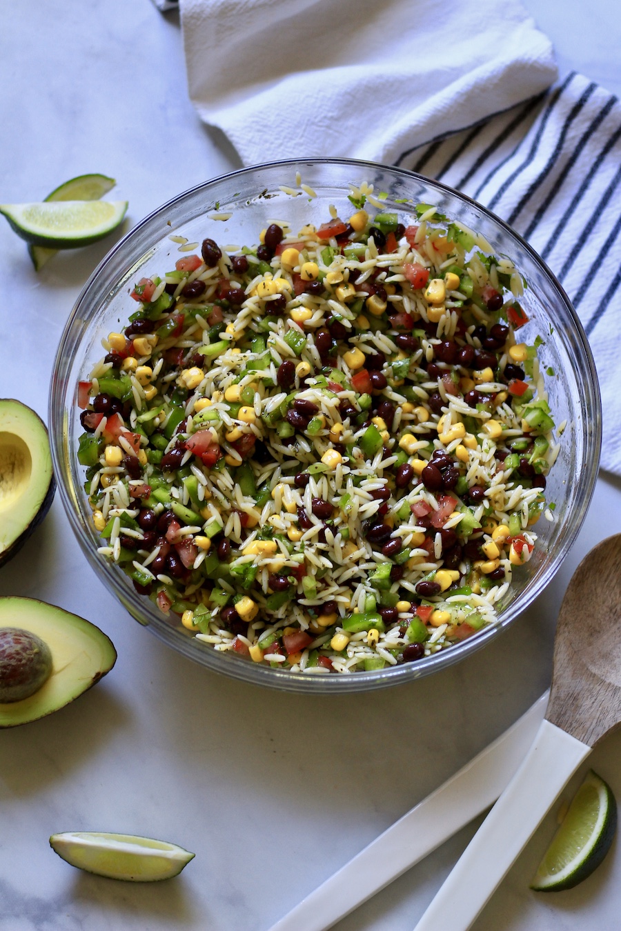 A glass bowl of Texas Caviar pasta salad with avocados to the left and serving spoons to the right.