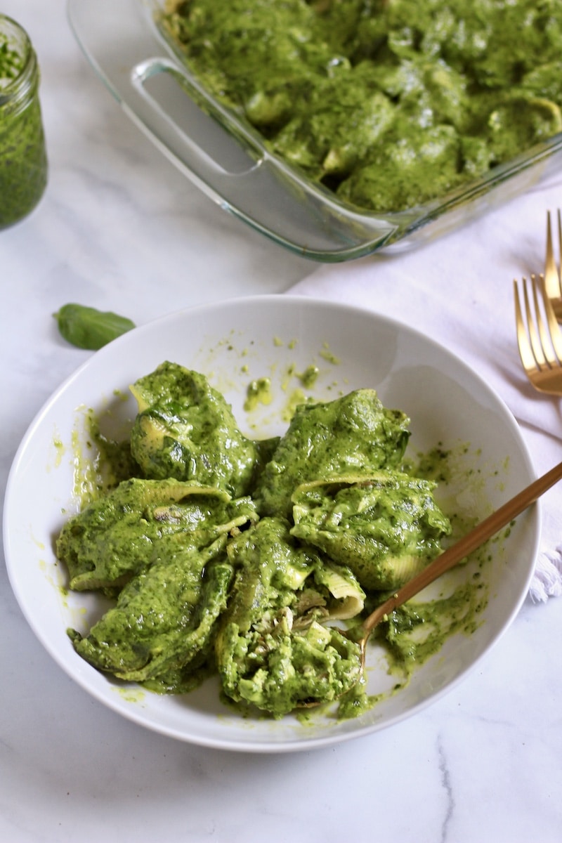 A white bowl of chicken pesto stuffed shells with a gold spoon and a baking dish in the back of baked chicken pesto stuffed shells.