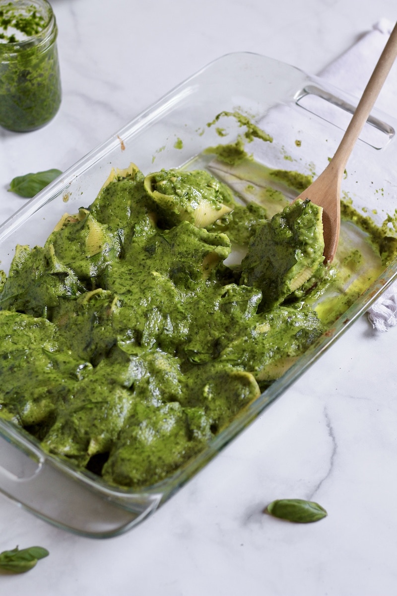 A glass baking dish with chicken pesto stuffed shells with a wooden spoon scooping one out.