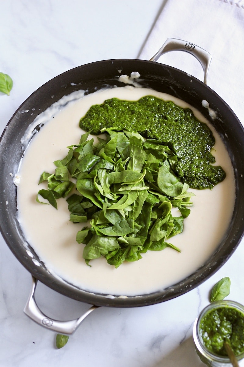 The pesto and spinach added to the base of the pesto cream sauce in a skillet on a white counter.