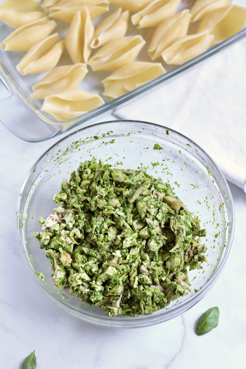 A glass bowl with shredded chicken tossed with pesto and a baking dish with large pasta shells in the back.