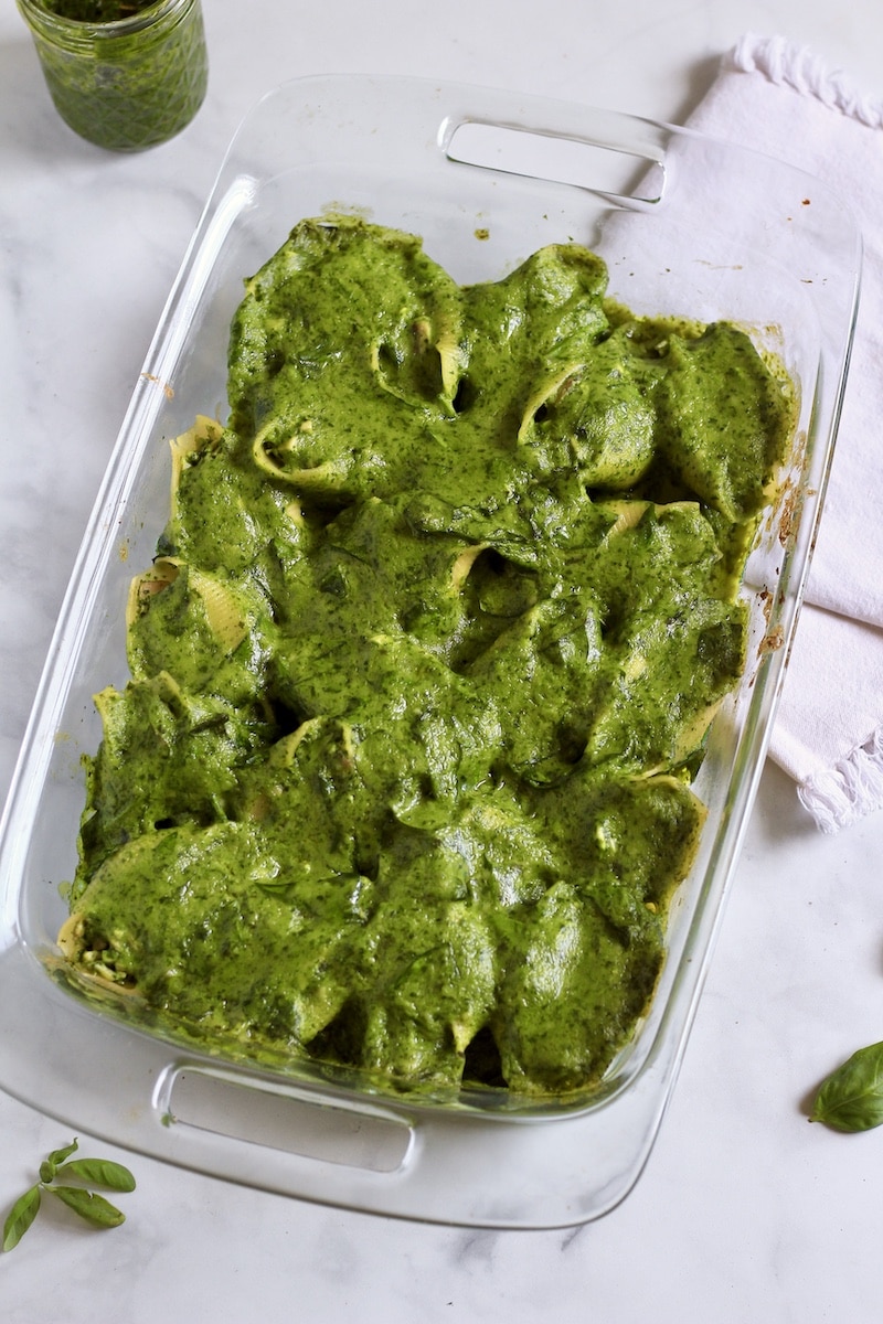 Spinach pesto cream sauce on top of chicken pesto stuffed shells in a baking dish on a white counter.