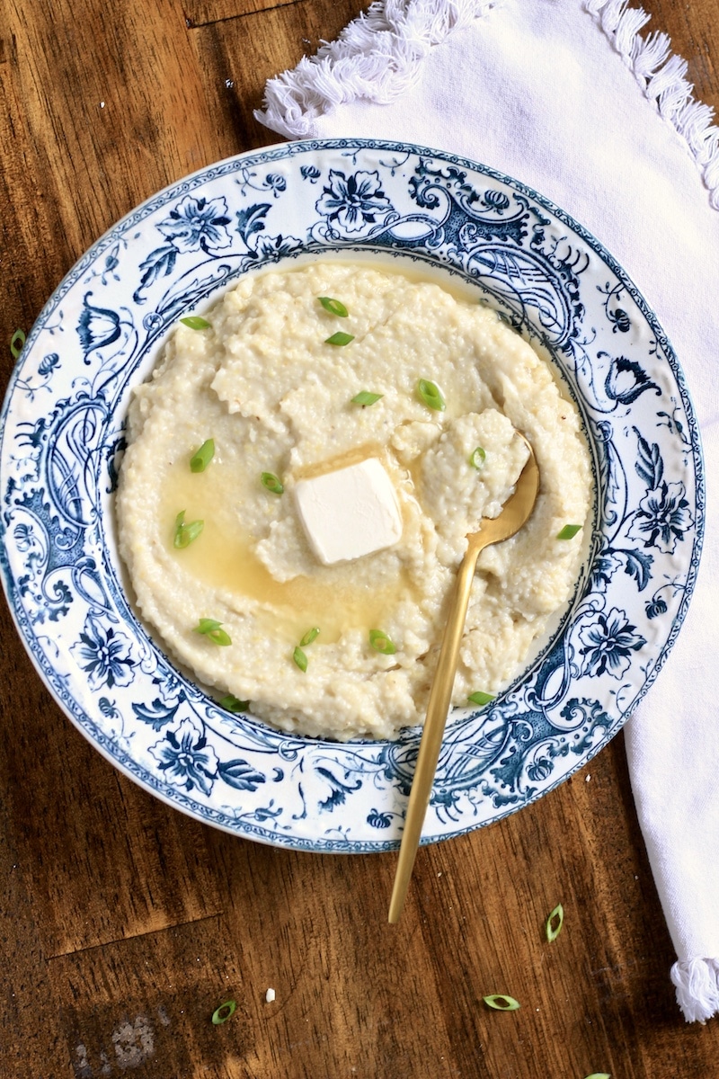 A top down photo of creamy grits in a blue and white rimmed bowl with a pad of vegan butter and green onion on top and a gold spoon in the bowl on the right.