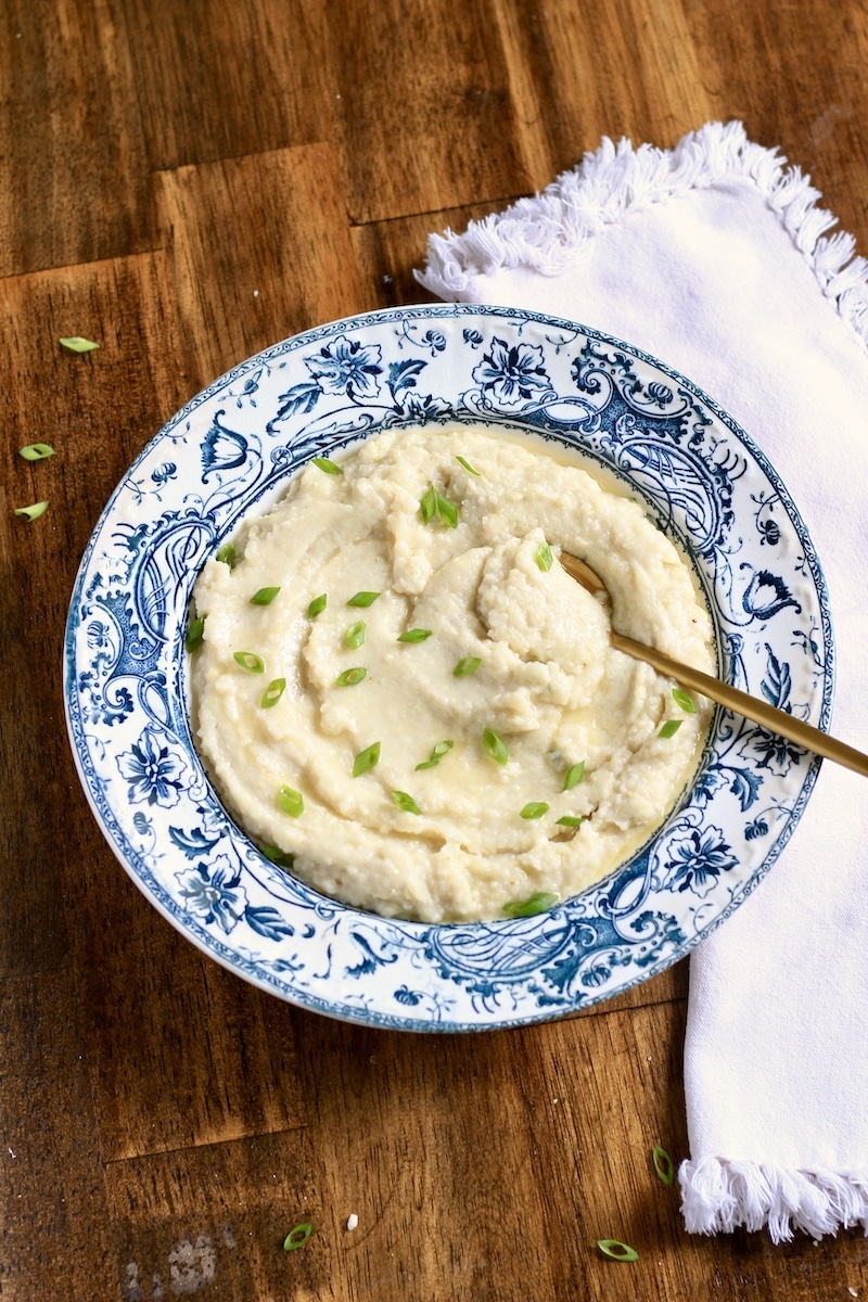 A blue and white rimmed bowl filled with creamy grits topped with green onion and a gold spoon to the top right and a white towel on the right side of the bowl.