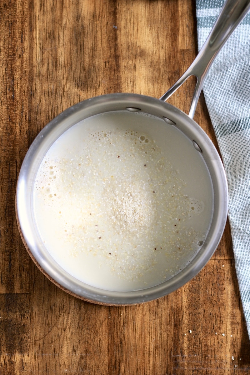 A silver saucepan on a wooden counter with boiled water and oat milk and grits added in.