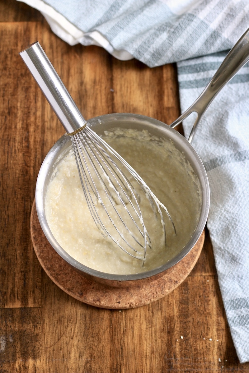 A top down photo of a silver saucepan with a wire whisk adding the grits into the liquid on a wooden counter.