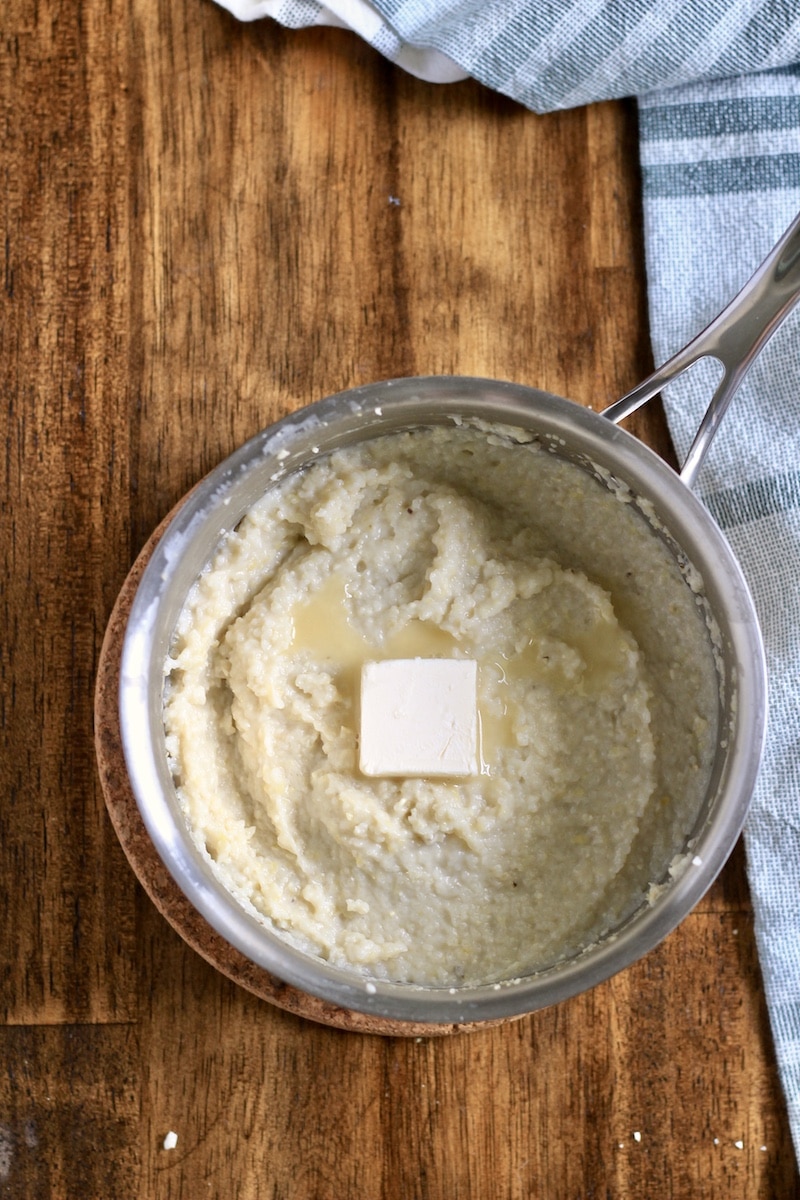 A silver saucepan with cooked creamy grits topped with a pad of butter on a wooden counter.