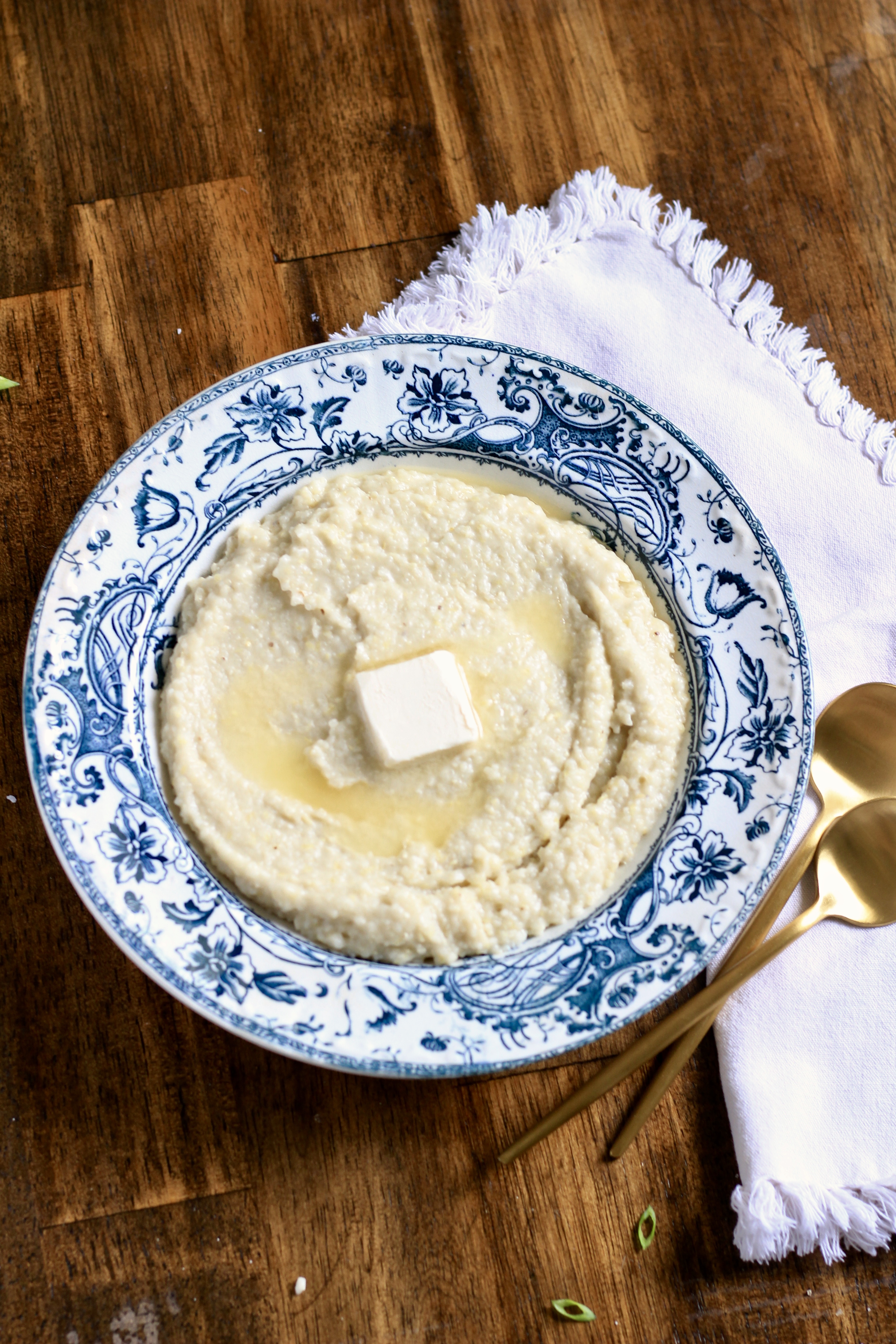 A blue and white bowl on a wooden counter with creamy grits topped with a pad of vegan butter with a white towel to the right with two golden spoons to the right.