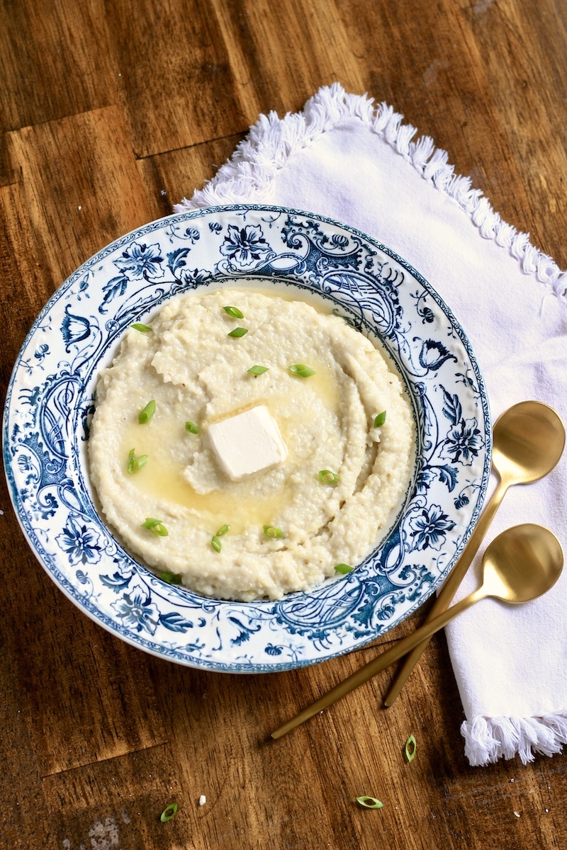 A blue and white rimmed bowl with creamy grits topped with green onion and vegan butter with two gold spoons to the right and a white towel.
