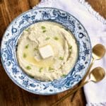 A square photo with a blue rimmed bowl next to two gold spoons and a serving of grits in the bowl topped with dairy-free butter.