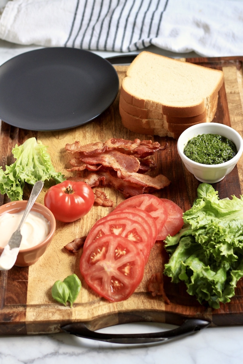 A wooden cutting board with the ingredients for pesto BLT and a blue and white striped dish towel in the back.