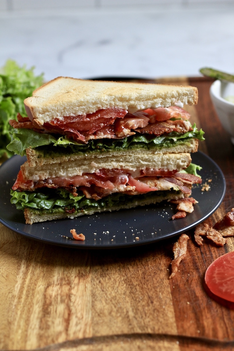 A plate with a stack of two triangular cut pesto blt sandwiches on a blue plate on top of a wooden cutting board with a slice of tomato to the front right.