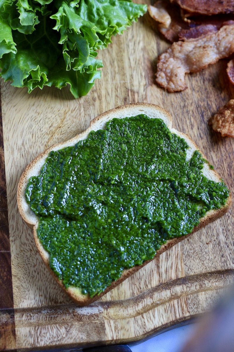 A slice of bread covered in pesto with lettuce and bacon above on a wooden cutting board.