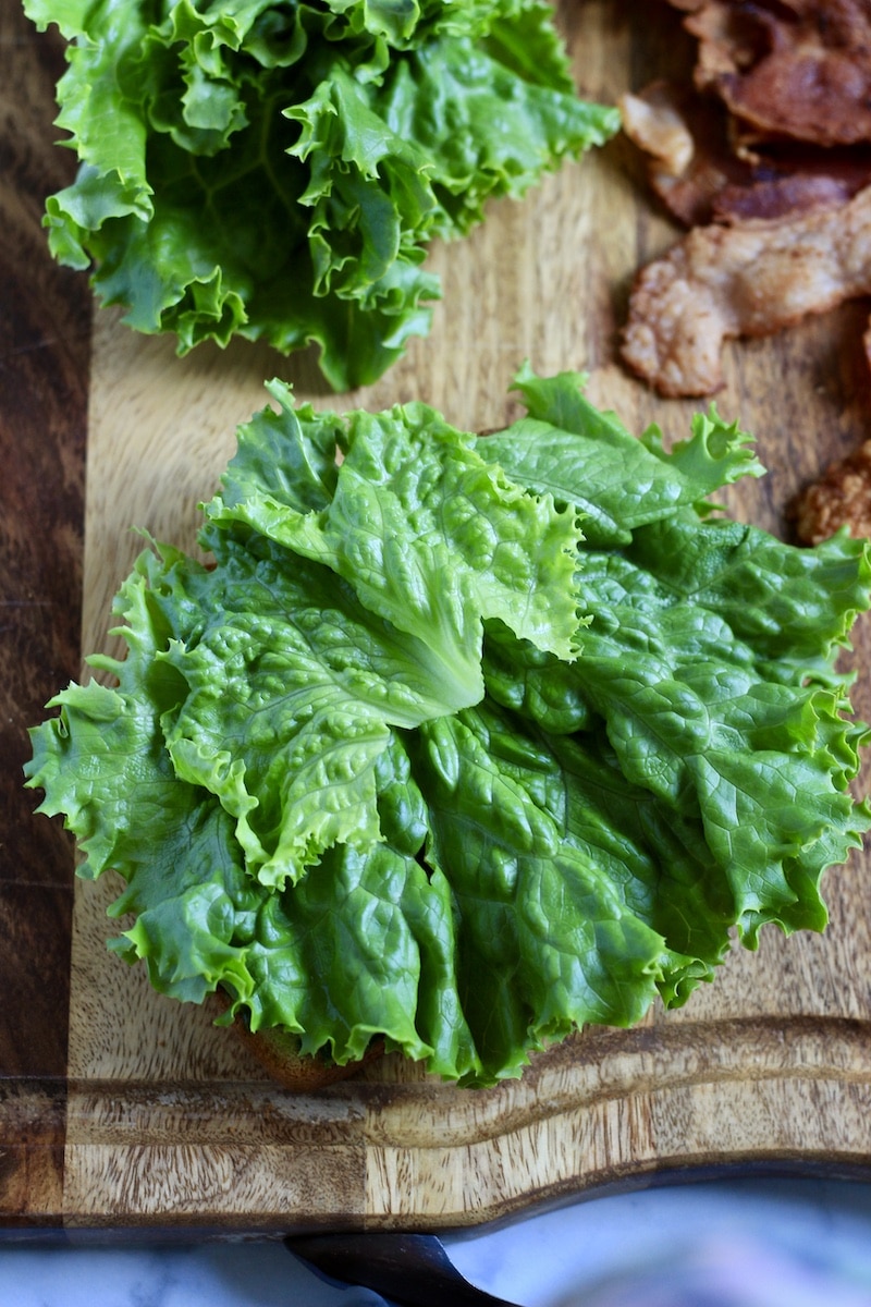 Lettuce on top of the pesto coated bread on a wooden cutting board.