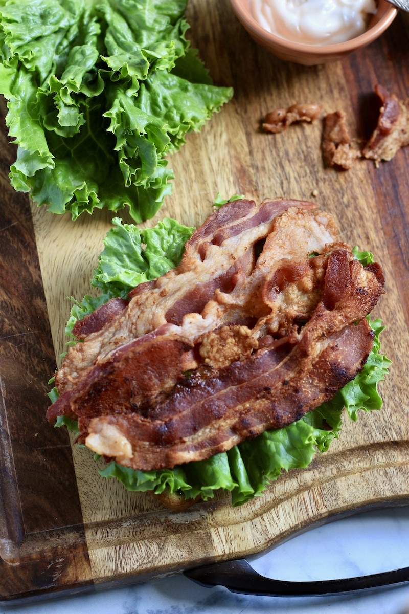 A top down photo with bacon on top of lettuce on a wooden cutting board with lettuce and a bowl of mayo in the back.