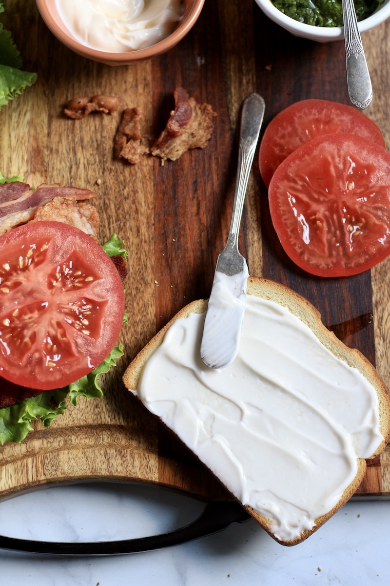 A slice of bread slathered with mayo with a silver knife on the right side of the cutting board and a stack of tomato slices behind it.