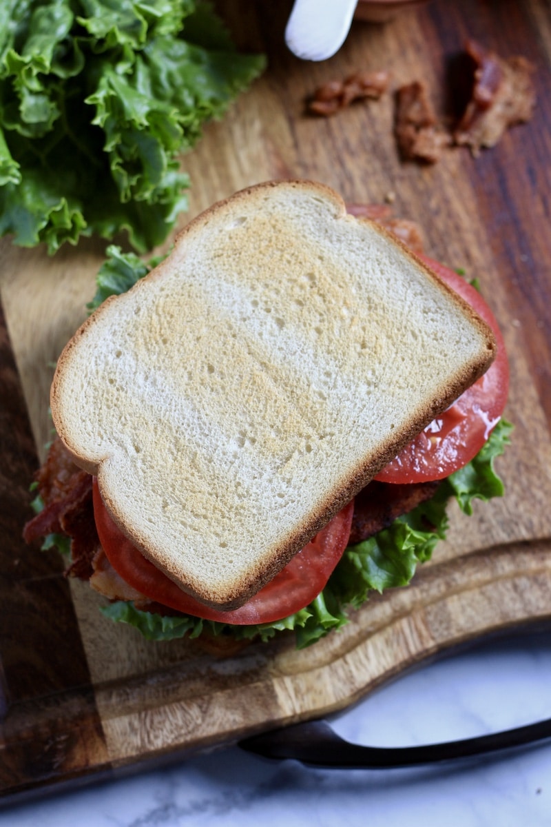 Top down photo of a pesto BLT on a wooden cutting board with lettuce in the back.