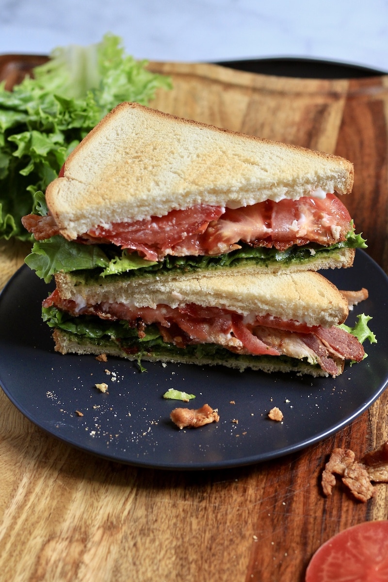 A toasted pesto BLT sandwich cut in two and stacked on top showing the cut side on a blue plate on a wooden counter.