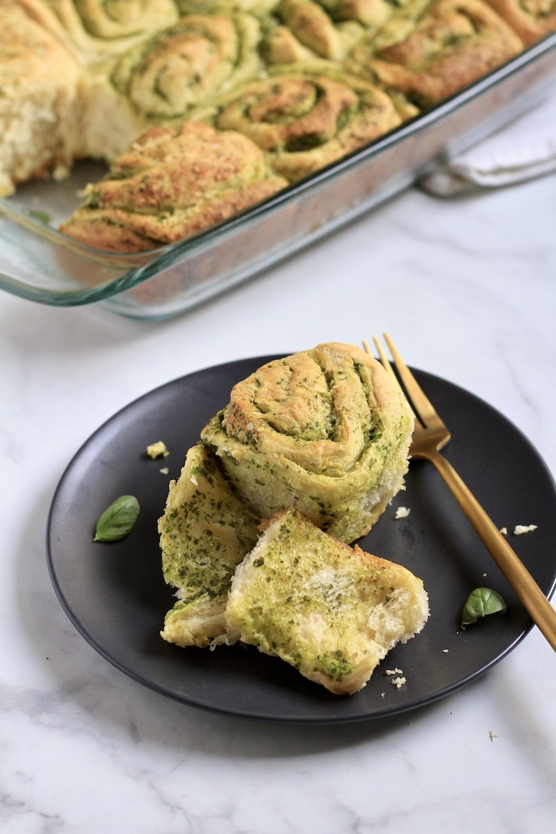A blue plate with a pesto roll with a gold fork and a baking dish of pesto rolls in the back.