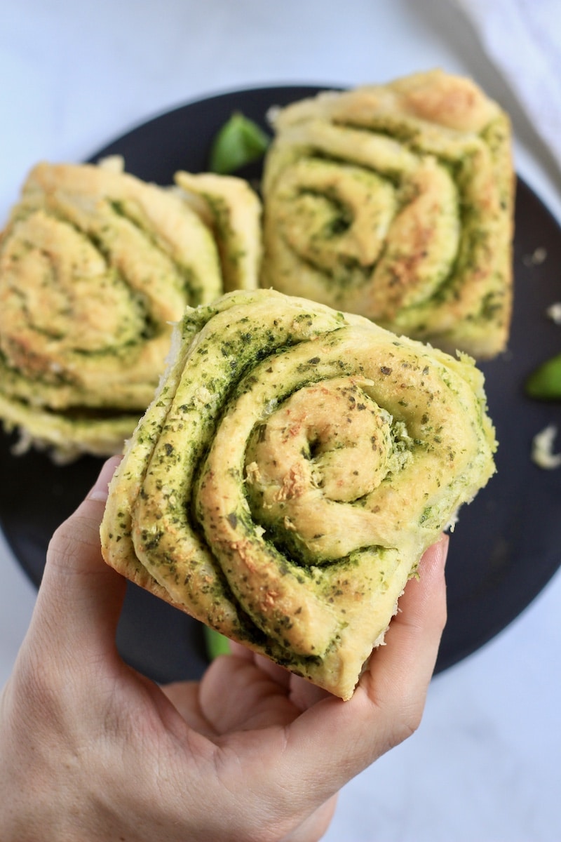 A hand holding a pesto roll in front of a plate with more pesto rolls.