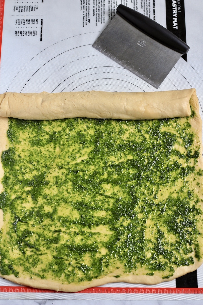 Pesto rolls dough with pesto spread on it and the dough being rolled from the back to the front.