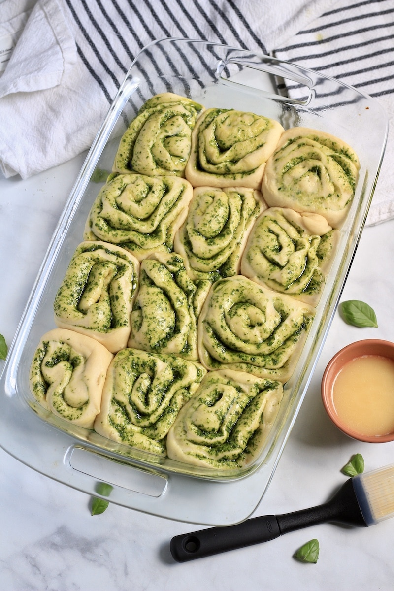 A glass baking dish with pesto rolls after rising with garlic butter and a pastry brush to the right.