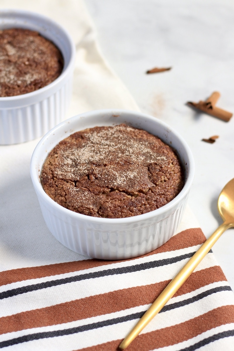A white ramekin with snickerdoodle baked oats on a towel in the center and a second baked oats in the back left with a gold spoon in the front right.