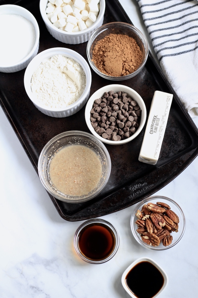 Ingredients for vegan brownies in small bowls on a dark cookie sheet with a blue and white striped towel to the back right.
