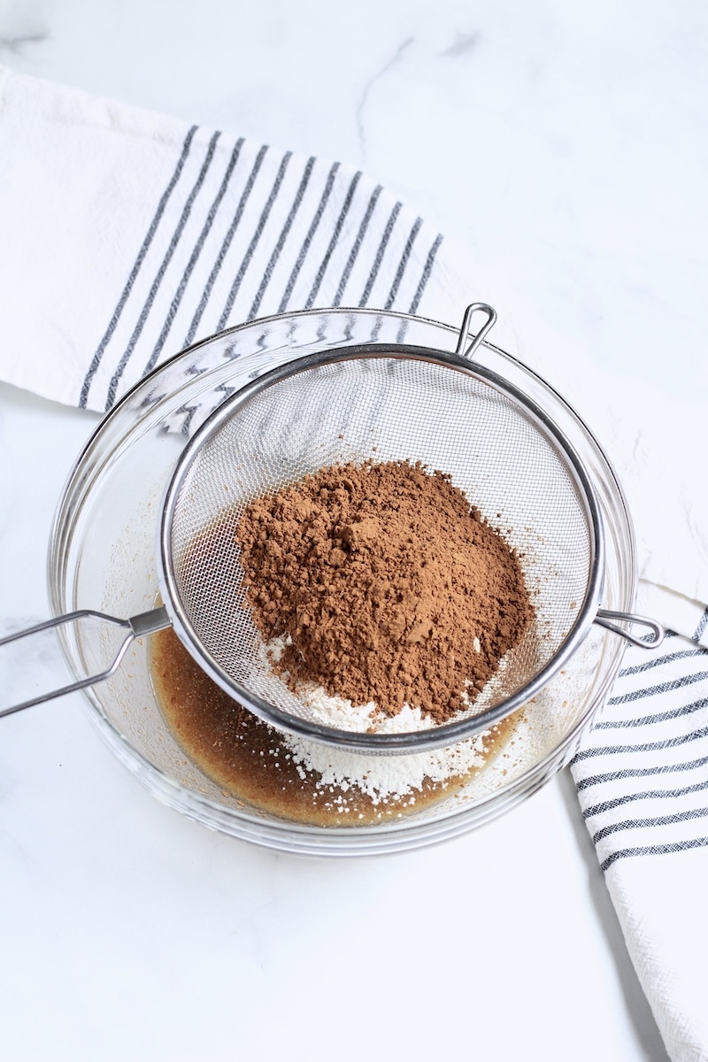 A glass bowl with a sifter on top and cocoa powder sifting into the glass bowl on bottom with a white and blue striped dish towel in the back.