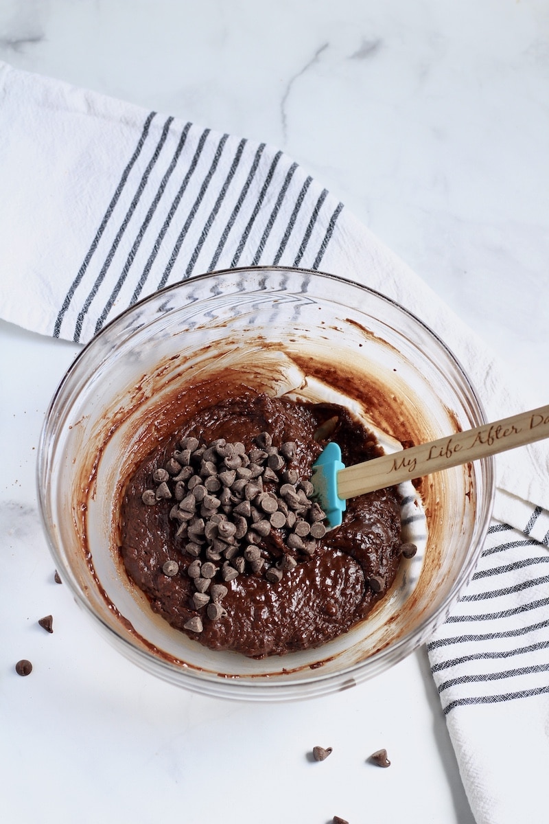 A glass bowl with vegan brownie batter and some chocolate chips on top with a spatula in the right side of the bowl.