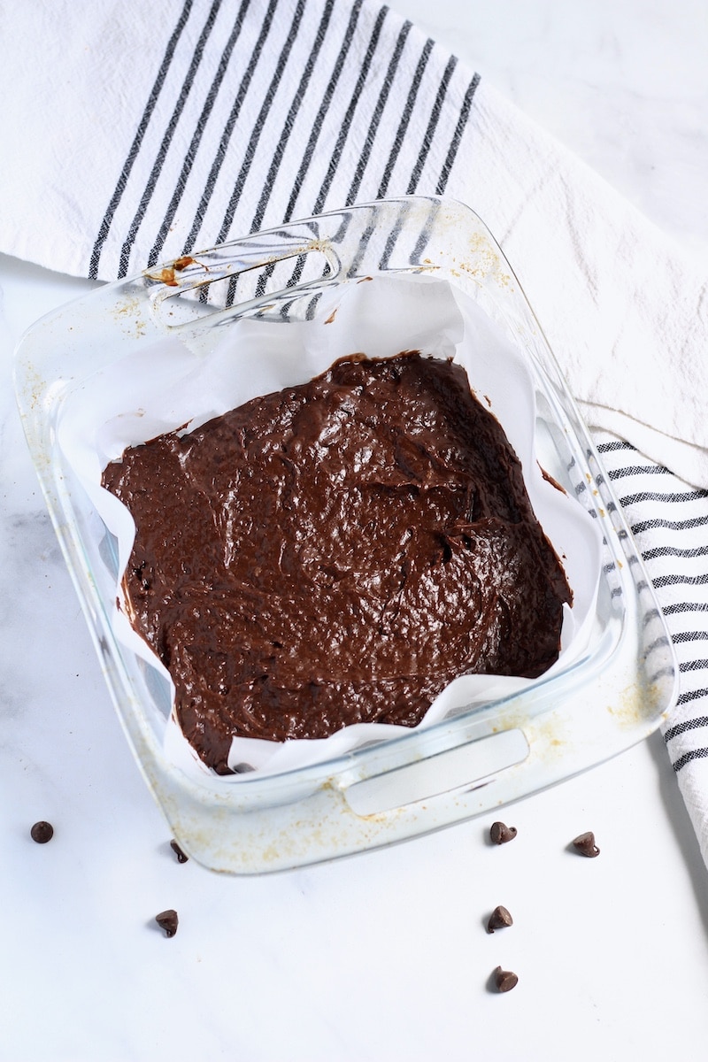 A square glass baking dish lined with parchment paper and filled with vegan brownie batter.