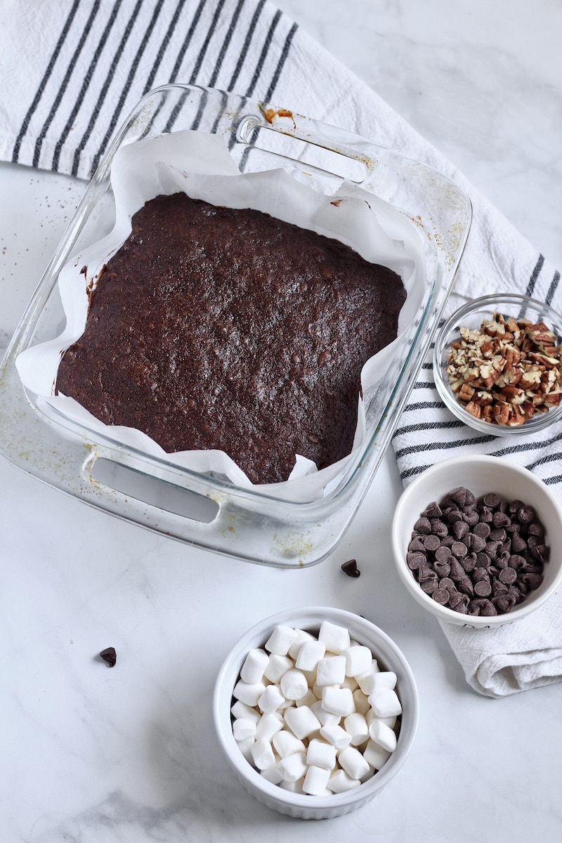 Vegan brownies in a glass baking pan with rocky road toppings in small bowls around the pan of brownies.