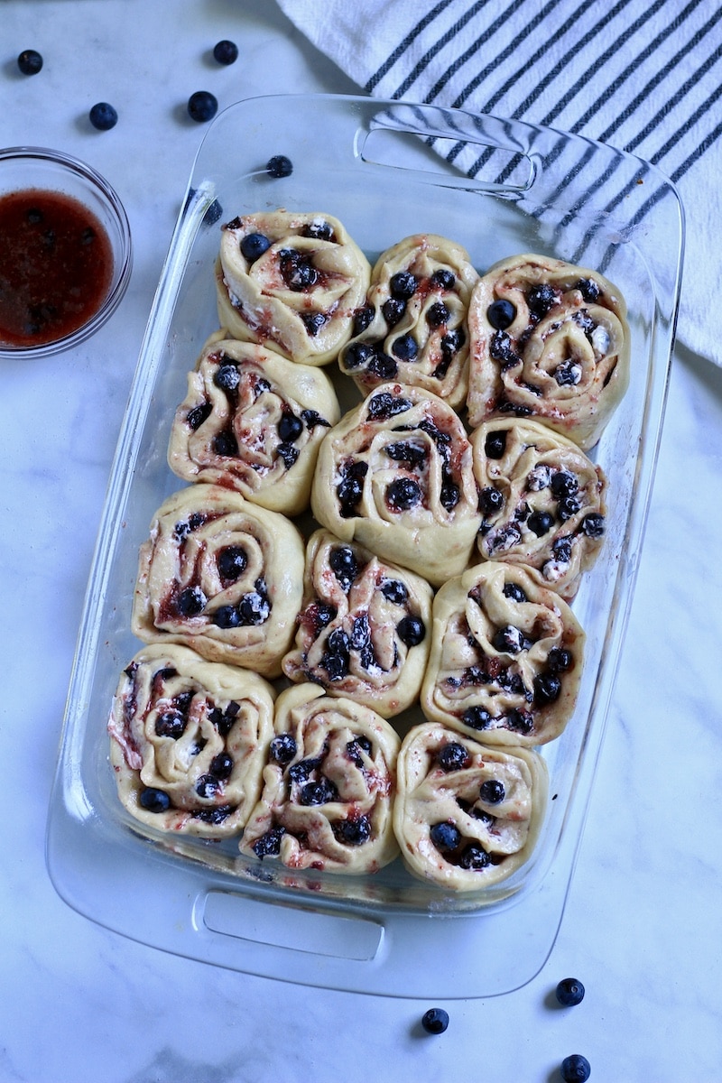 A glass baking dish with sweet rolls filled with lemon and blueberry on a counter with a blue and white striped dish towel to the top right.